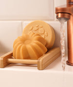 Lamazuna Keratin Shine solid conditioner bar resting in a wooden soap dish with the Keratin Repair shampoo bar behind it beside a running tap
