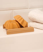 Lamazuna Keratin Shine solid conditioner bar in a wooden soap dish next to folded white towels in a bathroom setting
