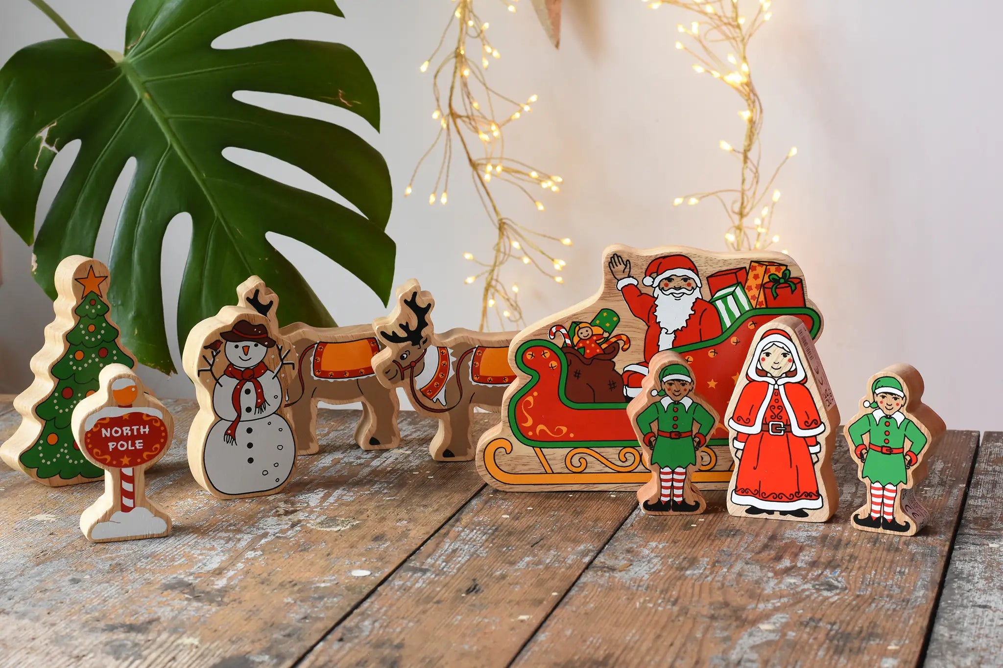 Lanka Kade wooden Christmas play set. 9 toy figures including Santa in his sleigh, Mrs Clause, Christmas Tree, Snowman, North Pole Sign, 2 Elves and 2 Reindeer placed on a wooden surface in the Babipur playroom. 