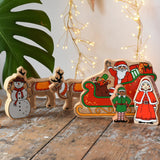 Lanka Kade wooden Christmas play set. 9 toy figures including Santa in his sleigh, Mrs Clause, Christmas Tree, Snowman, North Pole Sign, 2 Elves and 2 Reindeer placed on a wooden surface in the Babipur playroom. 