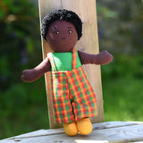 A Lanka Kade Boy Doll with Black Skin and Black Hair leaning against a wooden post outdoors.