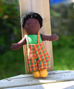 A Lanka Kade Boy Doll with Black Skin and Black Hair leaning against a wooden post outdoors.