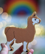 A Lanka Kade brown and white Llama wooden toy figure in an adult's hand. These wooden toy figures are part of a huge range of Lanka Kade fair trade, wooden toys available here at Babipur.