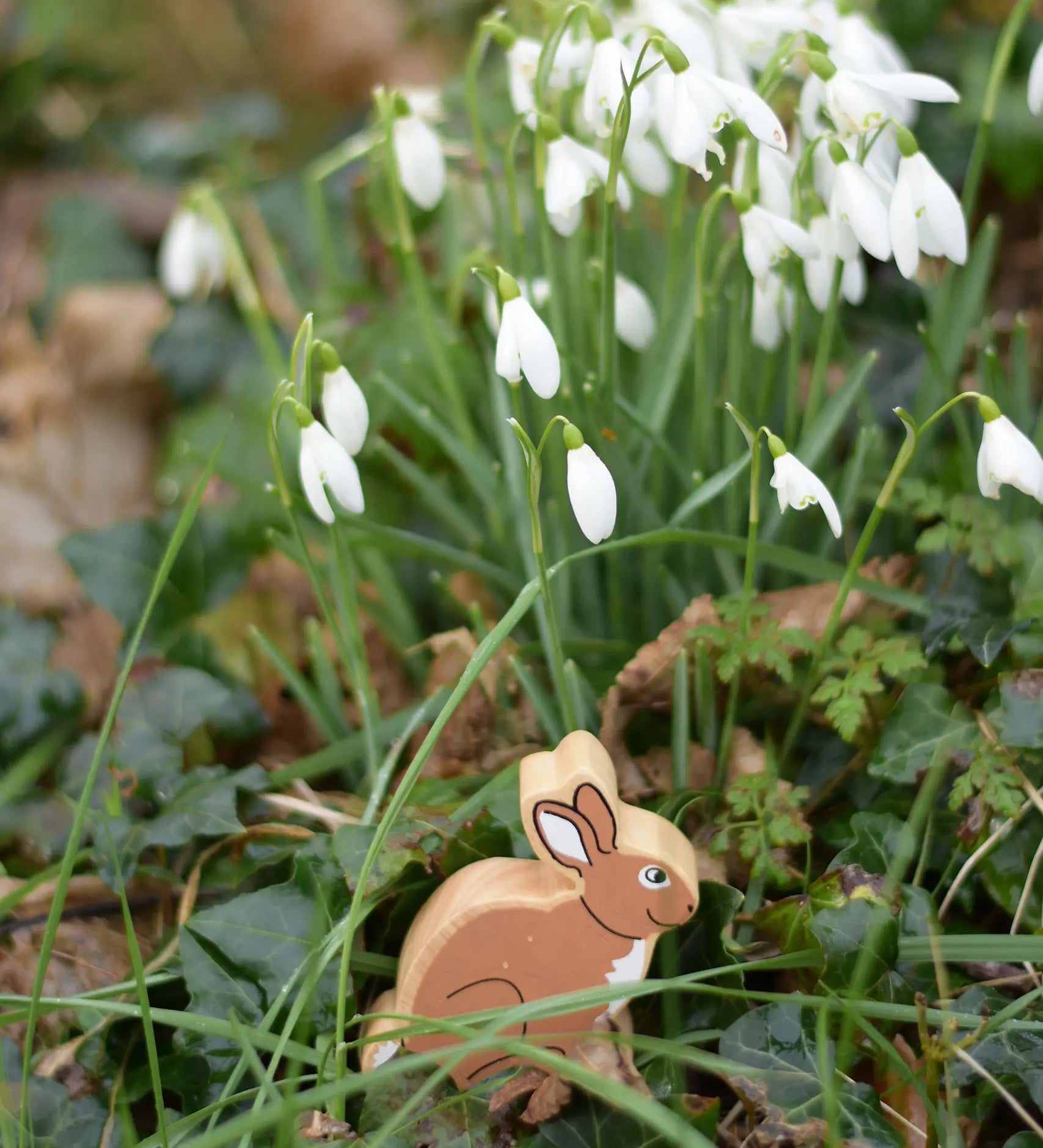 Lanka Kade brown bunny rabbit wooden toy figure placed on grass in front of a bunch of snowdrops. 