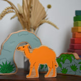 A Lanka Kade camel wooden toy figure placed on a small wooden table with the scenery pieces from the prehistoric play set. A Grimm's Wankel stacking toy can be seen in the background. These wooden toy figures are part of a huge range of Lanka Kade fair trade, wooden toys available here at Babipur.
