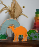 A Lanka Kade camel wooden toy figure placed on a small wooden table with the scenery pieces from the prehistoric play set. A Grimm's Wankel stacking toy can be seen in the background. These wooden toy figures are part of a huge range of Lanka Kade fair trade, wooden toys available here at Babipur.