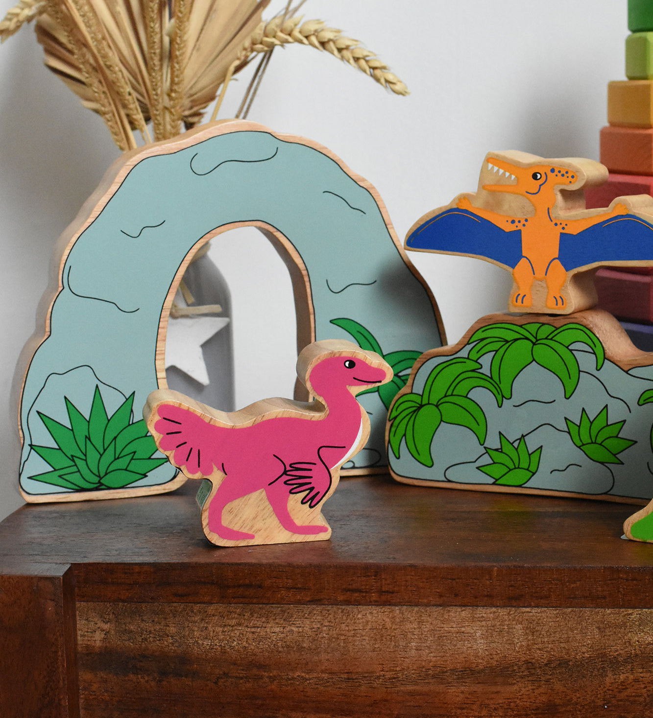 A Lanka Kade caudipteryx pink dinosaur wooden toy figure placed on a small wooden table with scenery pieces from the prehistoric play set in the background. These wooden toy figures are part of a huge range of Lanka Kade fair trade, wooden toys available here at Babipur.