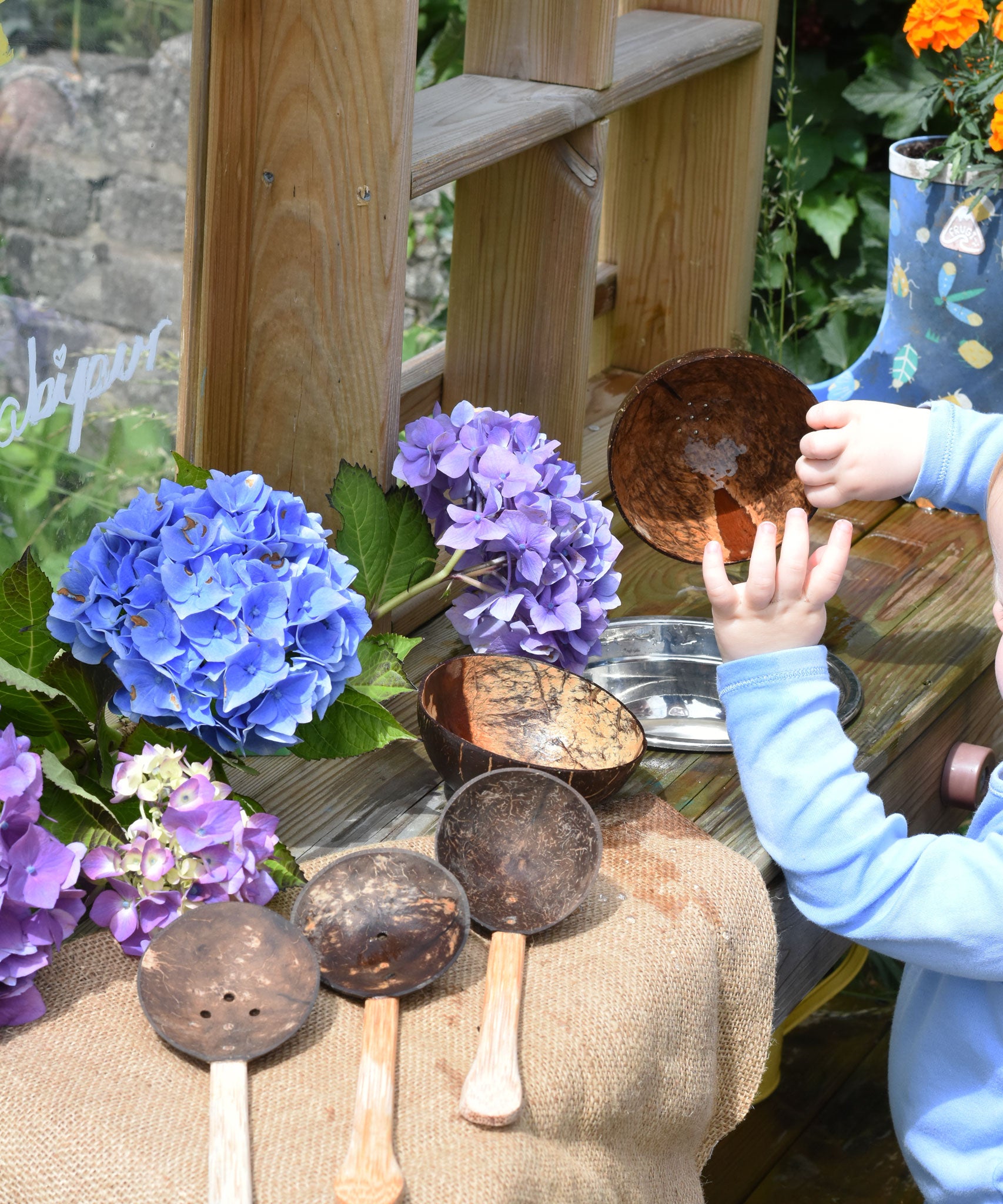 The Lanka Kade coconut shell water playset placed in a mud kitchen. There is a child playing with a coconut shell bowl, placing and lifting it out of a metal bowl that has been filled with water. 