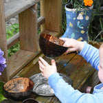 A child lifted the Lanka Kade coconut shell colander bowl from a metal bowl filled with water. Water can be seen dripping from the holes whilst the child touches the water. 