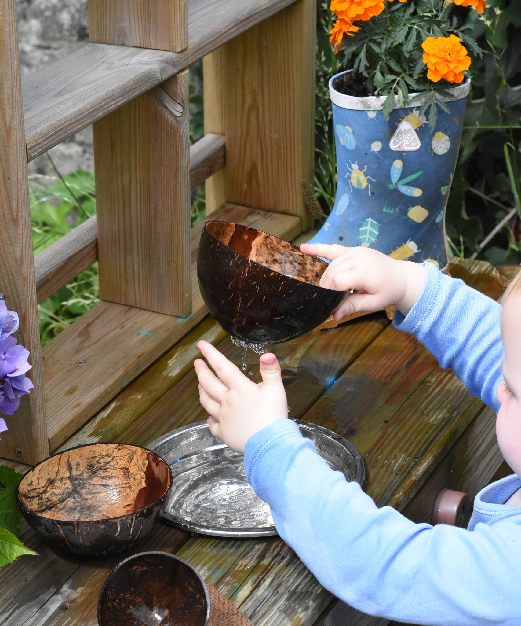 A child lifted the Lanka Kade coconut shell colander bowl from a metal bowl filled with water. Water can be seen dripping from the holes whilst the child touches the water. 