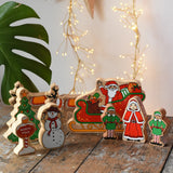 Lanka Kade wooden Christmas play set. 9 toy figures including Santa in his sleigh, Mrs Clause, Christmas Tree, Snowman, North Pole Sign, 2 Elves and 2 Reindeer placed on a wooden surface in the Babipur playroom. 