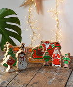 Lanka Kade wooden Christmas play set. 9 toy figures including Santa in his sleigh, Mrs Clause, Christmas Tree, Snowman, North Pole Sign, 2 Elves and 2 Reindeer placed on a wooden surface in the Babipur playroom. 