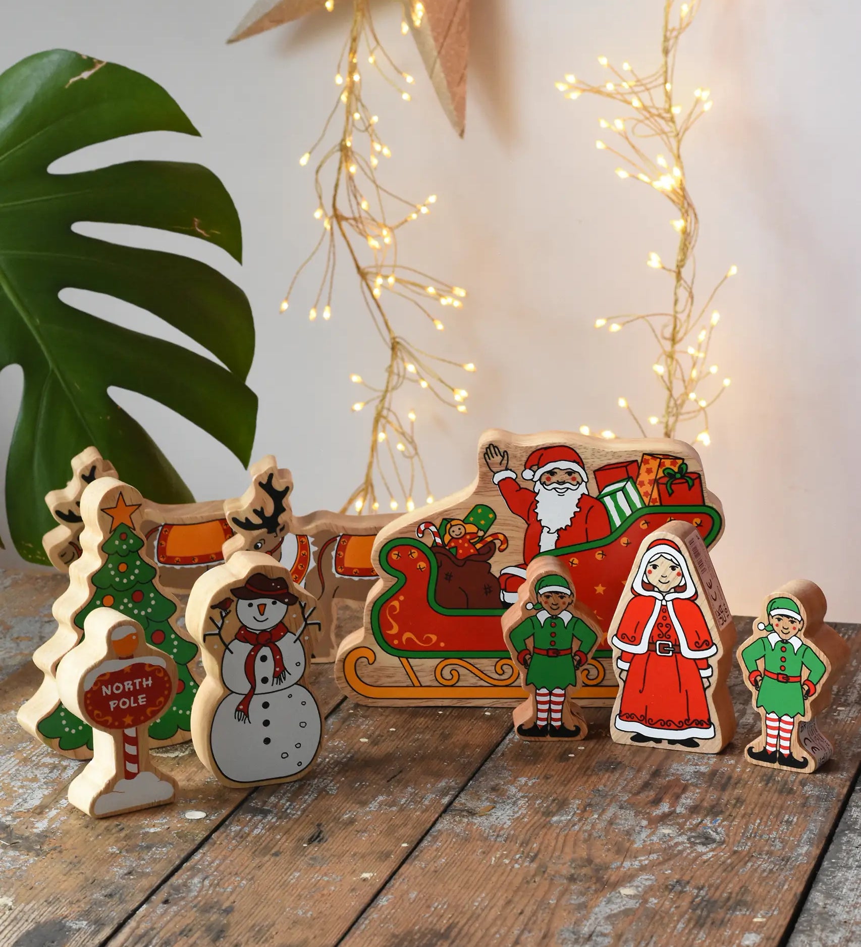 Lanka Kade wooden Christmas play set. 9 toy figures including Santa in his sleigh, Mrs Clause, Christmas Tree, Snowman, North Pole Sign, 2 Elves and 2 Reindeer placed on a wooden surface in the Babipur playroom. 