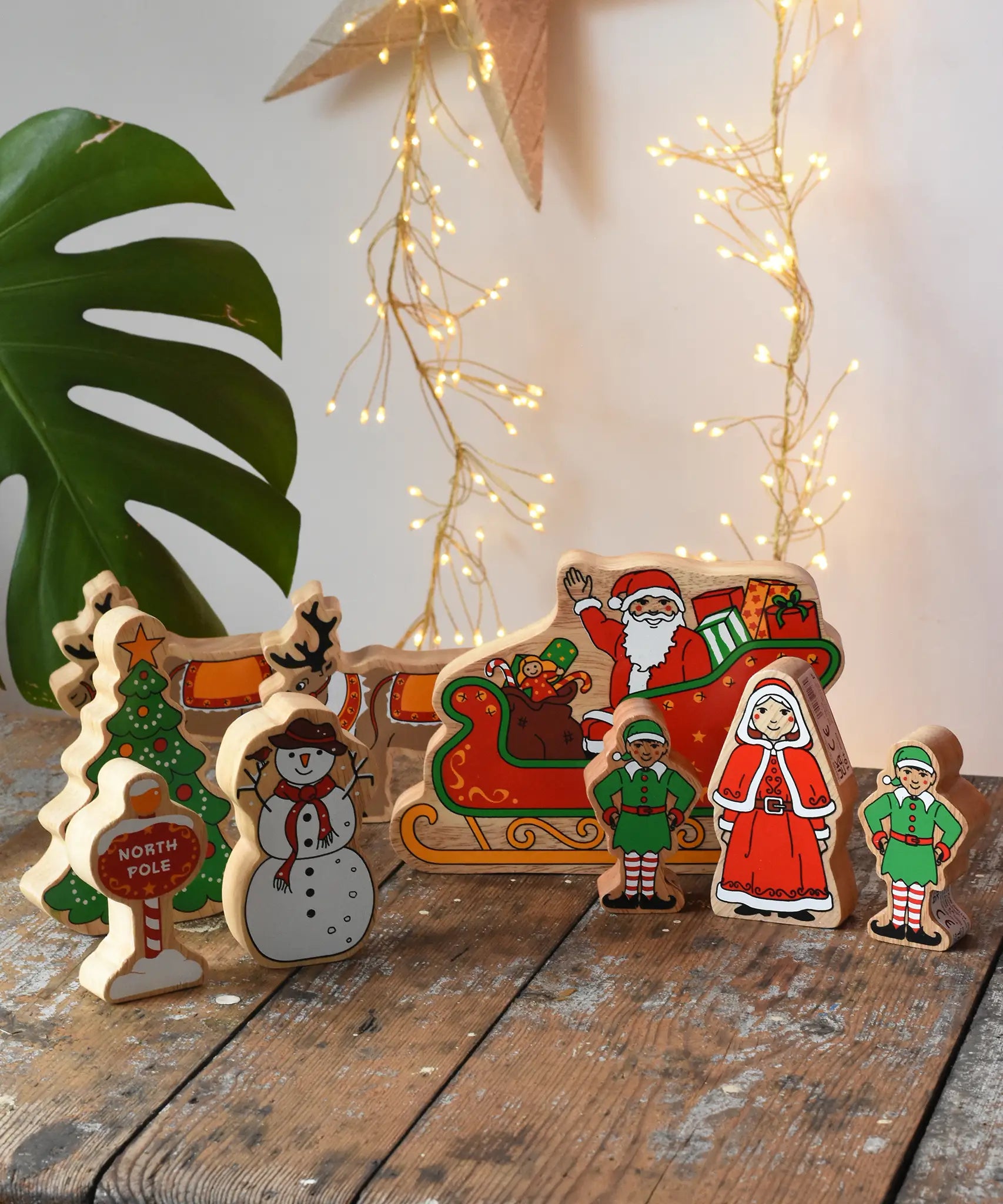 Lanka Kade wooden Christmas play set. 9 toy figures including Santa in his sleigh, Mrs Clause, Christmas Tree, Snowman, North Pole Sign, 2 Elves and 2 Reindeer placed on a wooden surface in the Babipur playroom. 