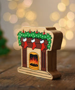 Lanka Kade festive fireplace wooden Christmas wooden toy figure. Brown fireplace with a lit fire, three red stocking hanging with a garland on the mantel decorated with 4 candles. 