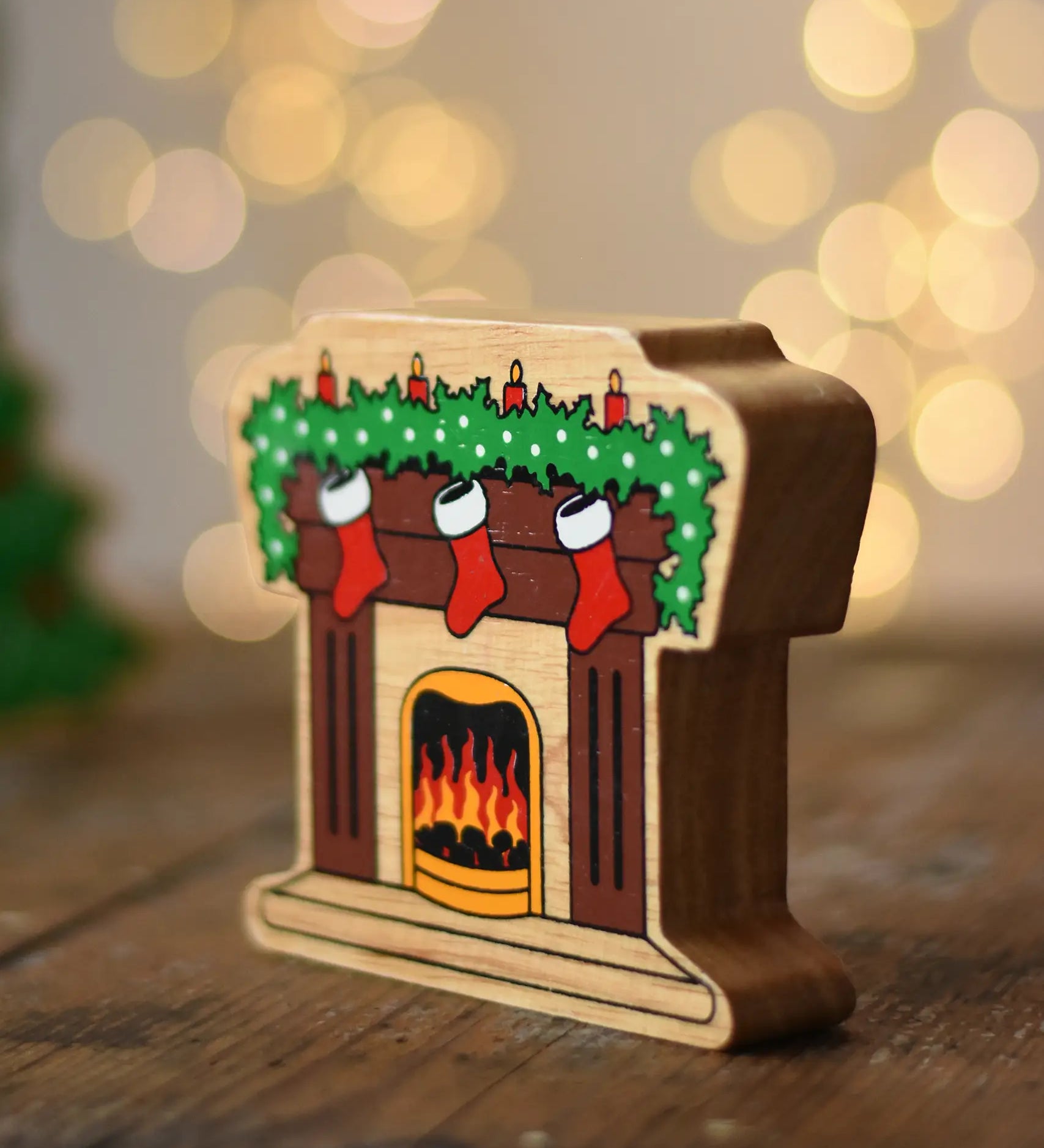 Lanka Kade festive fireplace wooden Christmas wooden toy figure. Brown fireplace with a lit fire, three red stocking hanging with a garland on the mantel decorated with 4 candles. 