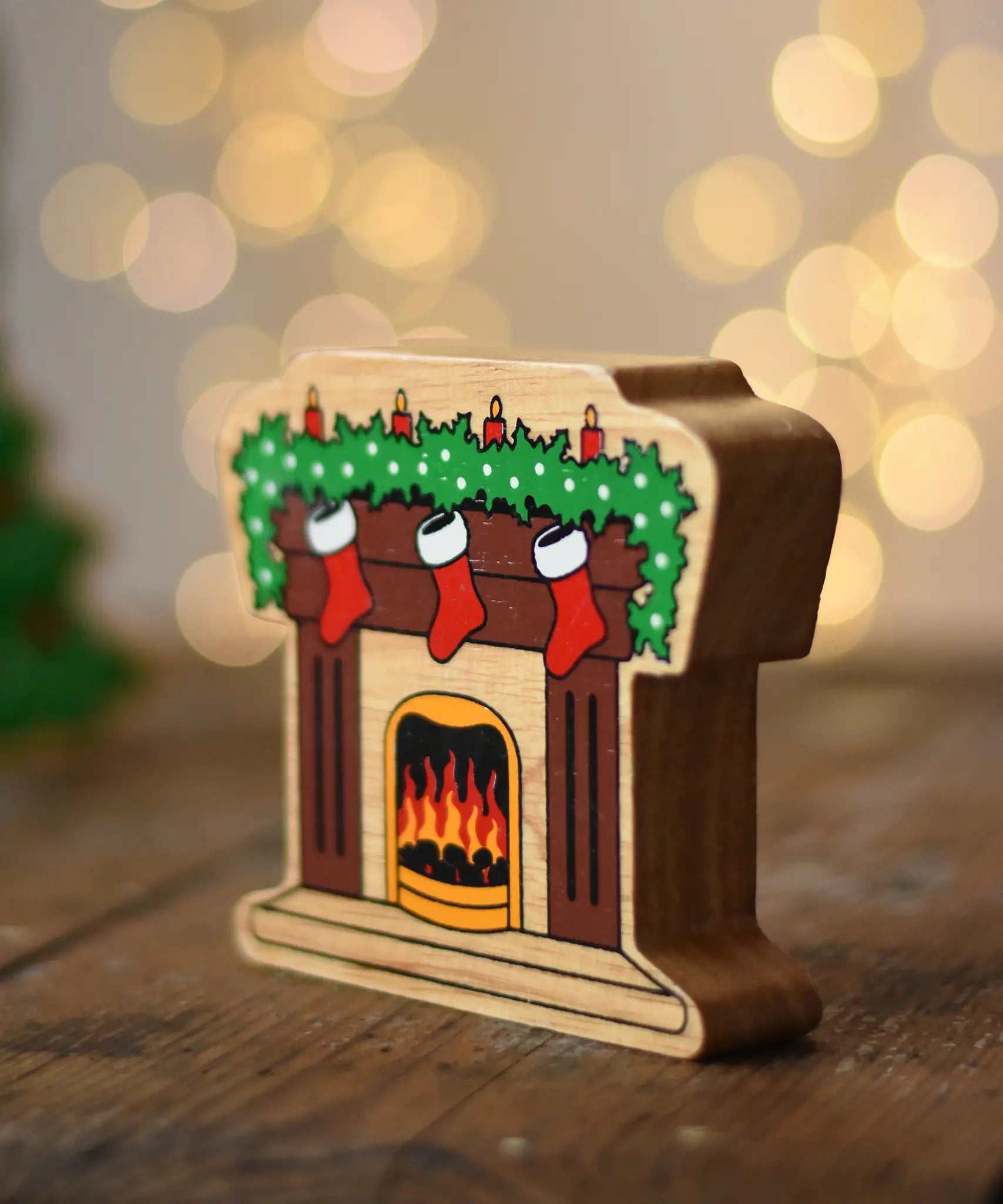 Lanka Kade festive fireplace wooden Christmas wooden toy figure. Brown fireplace with a lit fire, three red stocking hanging with a garland on the mantel decorated with 4 candles. 