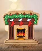 Lanka Kade festive fireplace wooden Christmas wooden toy figure. Brown fireplace with a lit fire, three red stocking hanging with a garland on the mantel decorated with 4 candles. 