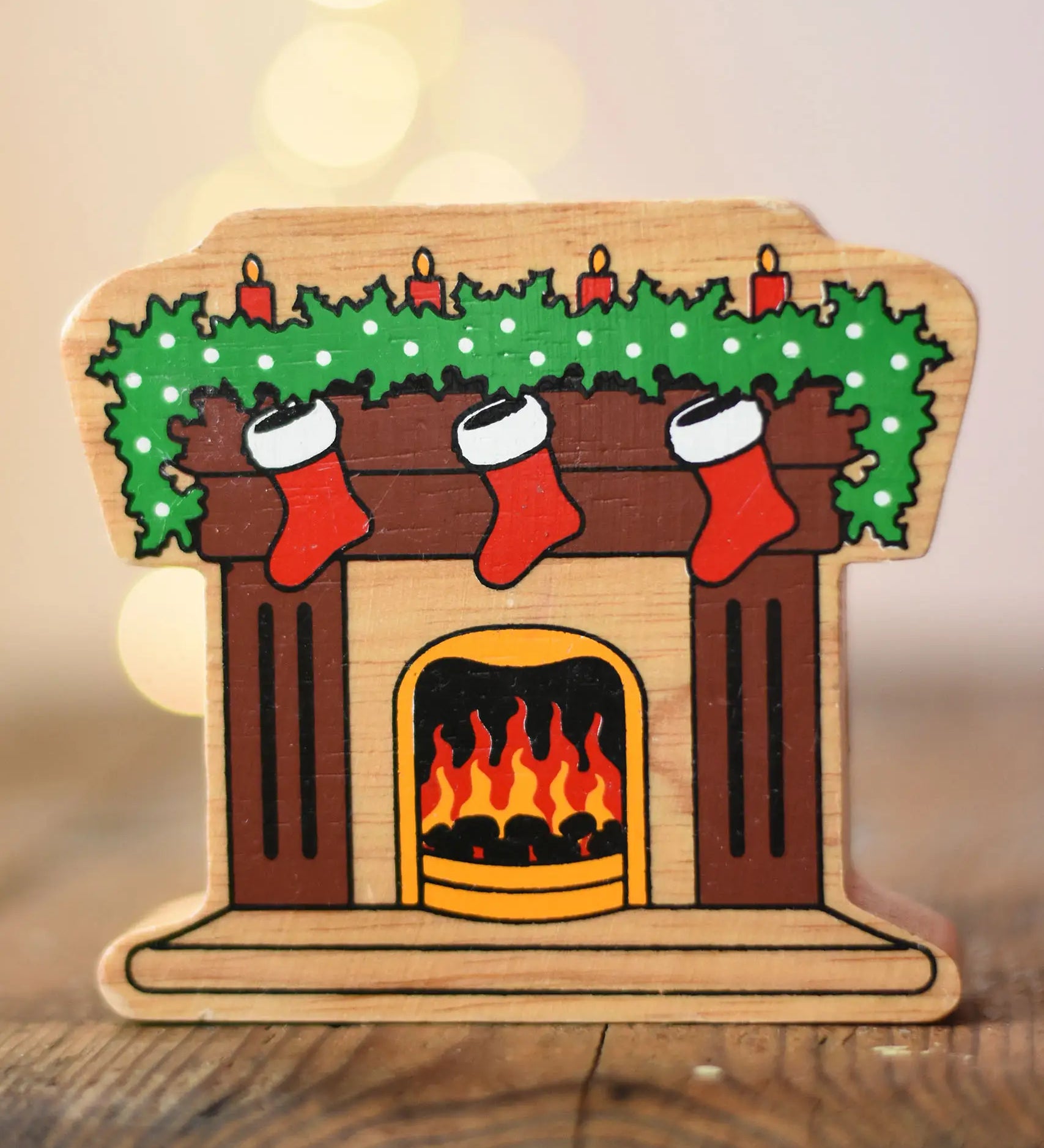 Lanka Kade festive fireplace wooden Christmas wooden toy figure. Brown fireplace with a lit fire, three red stocking hanging with a garland on the mantel decorated with 4 candles. 