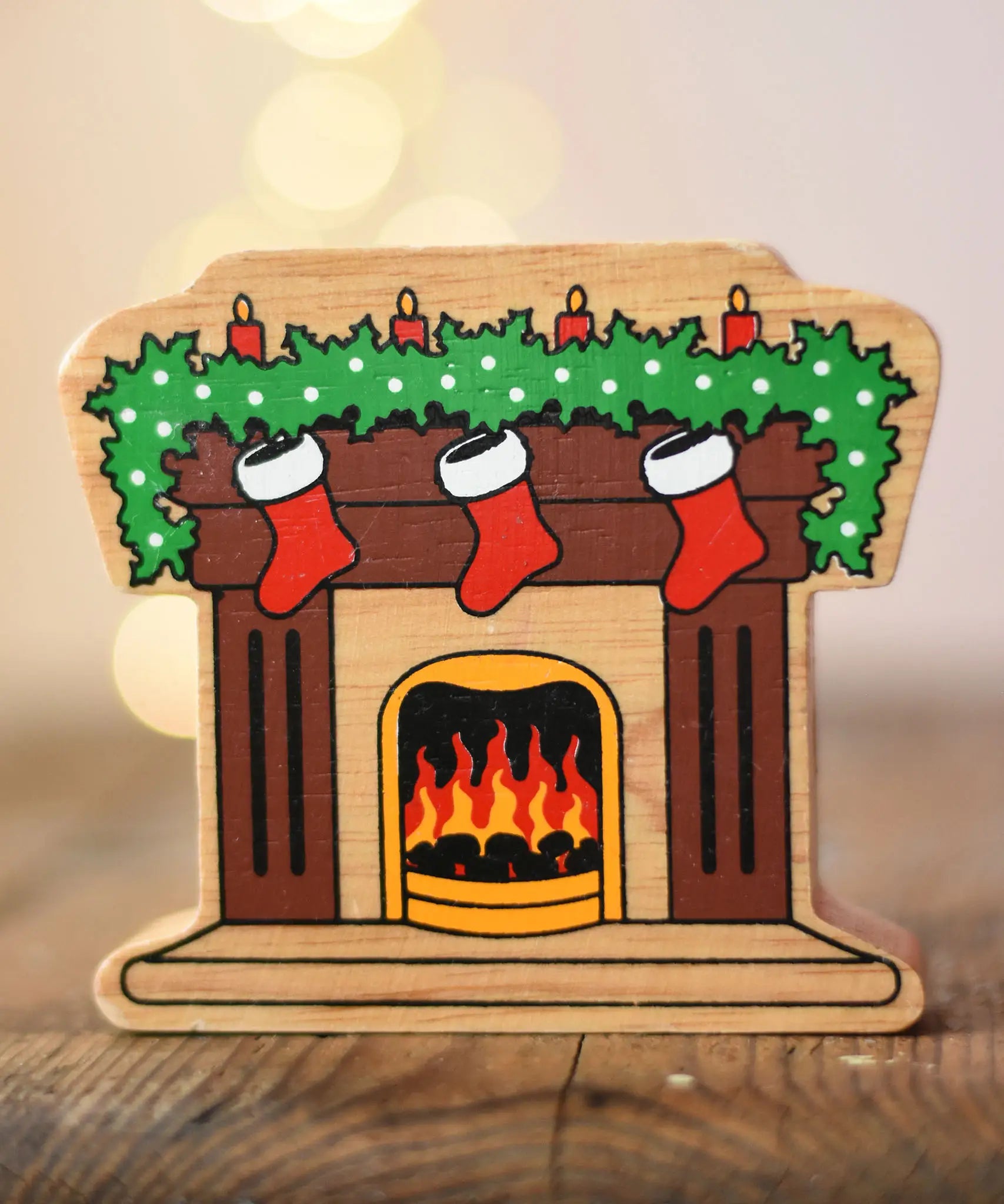 Lanka Kade festive fireplace wooden Christmas wooden toy figure. Brown fireplace with a lit fire, three red stocking hanging with a garland on the mantel decorated with 4 candles. 