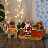 Lanka Kade wooden Christmas toy figures including Father Christmas,  red sleigh, Mrs Clause, North Pole Sign, Elf and 2 Reindeer placed on a wooden surface in the Babipur playroom.