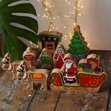 Lanka Kade wooden Christmas play set. 10 toy figures including Father Christmas,  red sleigh, Mrs Clause, Wrapped presents, festive fireplace, Christmas Tree, North Pole Sign, Elf and 2 Reindeer placed on a wooden surface in the Babipur playroom.