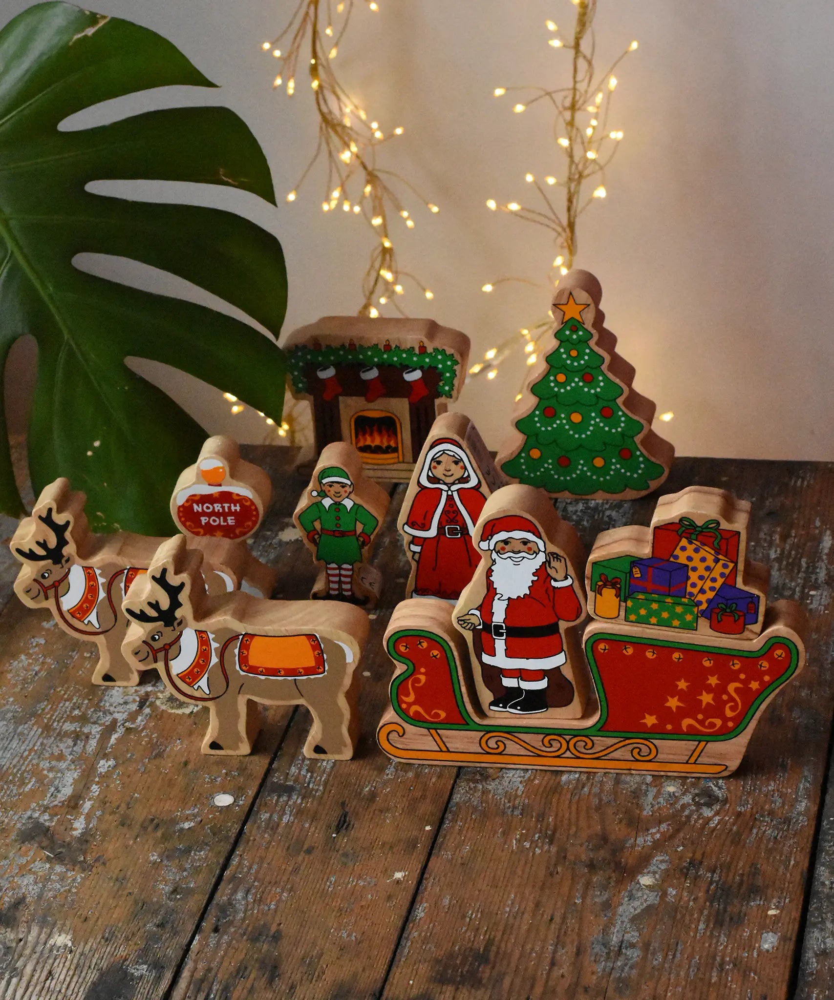 Lanka Kade wooden Christmas play set. 10 toy figures including Father Christmas,  red sleigh, Mrs Clause, Wrapped presents, festive fireplace, Christmas Tree, North Pole Sign, Elf and 2 Reindeer placed on a wooden surface in the Babipur playroom.