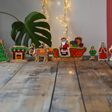 Lanka Kade wooden Christmas play set. 10 toy figures including Father Christmas,  red sleigh, Mrs Clause, Wrapped presents, festive fireplace, Christmas Tree, North Pole Sign, Elf and 2 Reindeer placed on a wooden surface in the Babipur playroom.