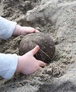 A child placing an upturned Lanka Kade coconut bowl on the sand at a beach. 