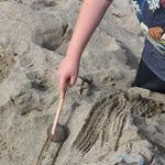 A child using the coconut rake utensil to make marks on the sand at a beach. The rake is part of a collection of coconut tools and bowls from Lanka Kade. 
