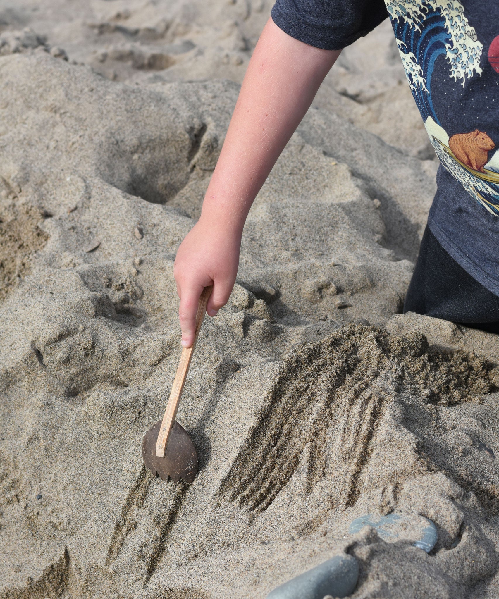 A child using the coconut rake utensil to make marks on the sand at a beach. The rake is part of a collection of coconut tools and bowls from Lanka Kade. 