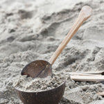 A rake utensil placed in a coconut bowl filled with sand on a beach. Other coconut utensils from the Lanka Kade coconut range can be seen in the background.
