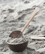 A rake utensil placed in a coconut bowl filled with sand on a beach. Other coconut utensils from the Lanka Kade coconut range can be seen in the background.