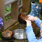 A close up of a child lifting the Lanka Kade colander coconut bowl from a bowl of water in a mud kitchen. Water can be seen steaming from the holes in the bottom of the coconut shell.