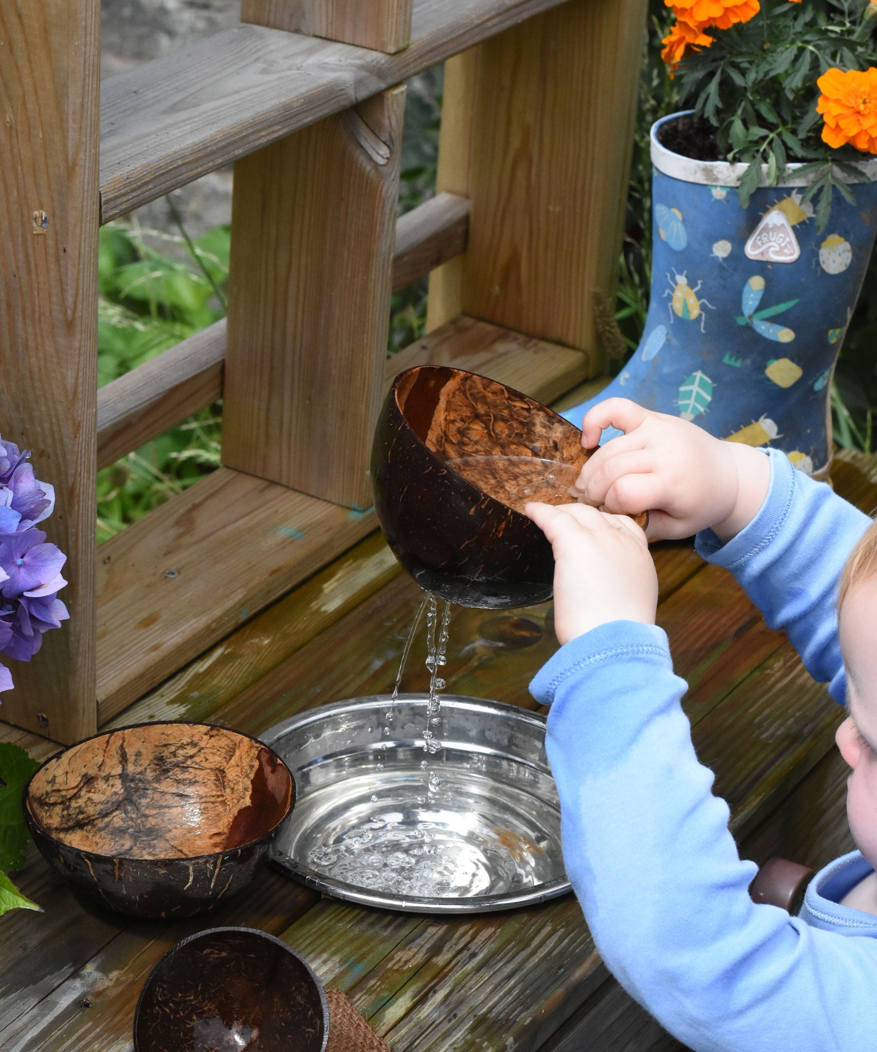 A close up of a child lifting the Lanka Kade colander coconut bowl from a bowl of water in a mud kitchen. Water can be seen steaming from the holes in the bottom of the coconut shell.