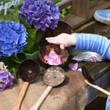 A close up of a child's hand placing pink coloured peony petals into a Lanka Kade coconut shell bowl. The the bowl and utensils from the water play set can be seen placed around the mud kitchen. 