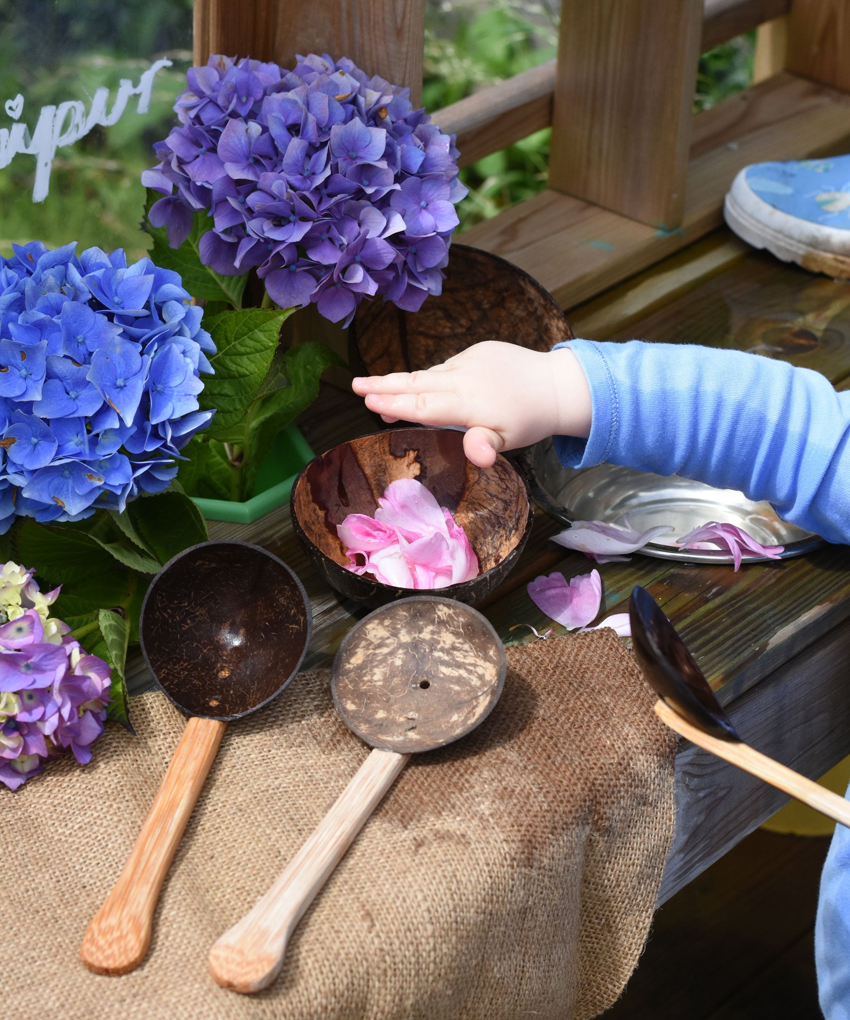 A close up of a child's hand placing pink coloured peony petals into a Lanka Kade coconut shell bowl. The the bowl and utensils from the water play set can be seen placed around the mud kitchen. 