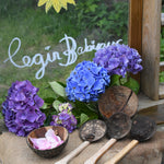 The Lanka Kade Coconut Shell Water Playset laid out on the surface of a mud kitchen outdoors. Blue and purple coloured hydrangeas have been placed behind them and the words 'Cegin Babipur' which is welsh for Babipur Kitchen written with Kitpas crayons on a perspex board behind them. 