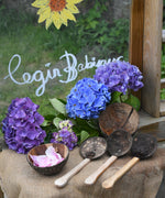 The Lanka Kade Coconut Shell Water Playset laid out on the surface of a mud kitchen outdoors. Blue and purple coloured hydrangeas have been placed behind them and the words 'Cegin Babipur' which is welsh for Babipur Kitchen written with Kitpas crayons on a perspex board behind them. 