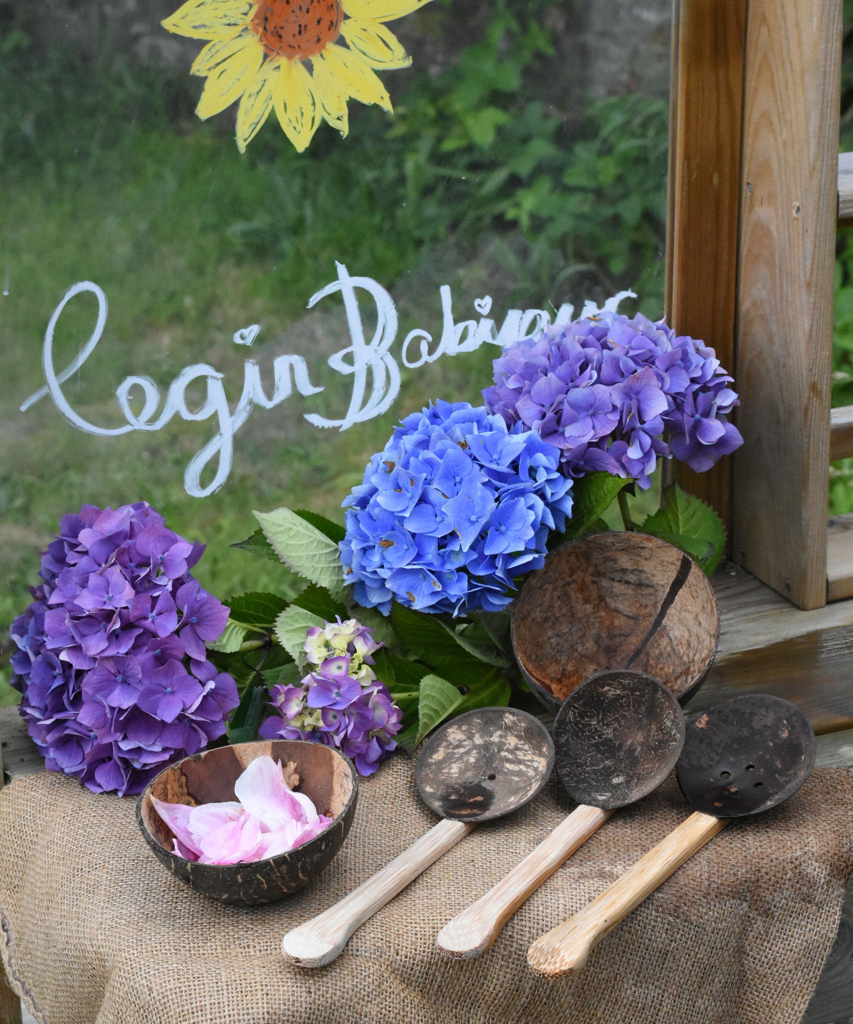 The Lanka Kade Coconut Shell Water Playset laid out on the surface of a mud kitchen outdoors. Blue and purple coloured hydrangeas have been placed behind them and the words 'Cegin Babipur' which is welsh for Babipur Kitchen written with Kitpas crayons on a perspex board behind them. 
