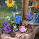 The Lanka Kade Coconut Shell Water Playset laid out on the surface of a mud kitchen outdoors. Blue and purple coloured hydrangeas have been placed behind them. A perspex board can be seen in the background with the words 'Cegin Babipur' which is welsh for Babipur Kitchen written with Kitpas crayons along with a drawing of a sunflower head.
