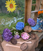 The Lanka Kade Coconut Shell Water Playset laid out on the surface of a mud kitchen outdoors. Blue and purple coloured hydrangeas have been placed behind them. A perspex board can be seen in the background with the words 'Cegin Babipur' which is welsh for Babipur Kitchen written with Kitpas crayons along with a drawing of a sunflower head.