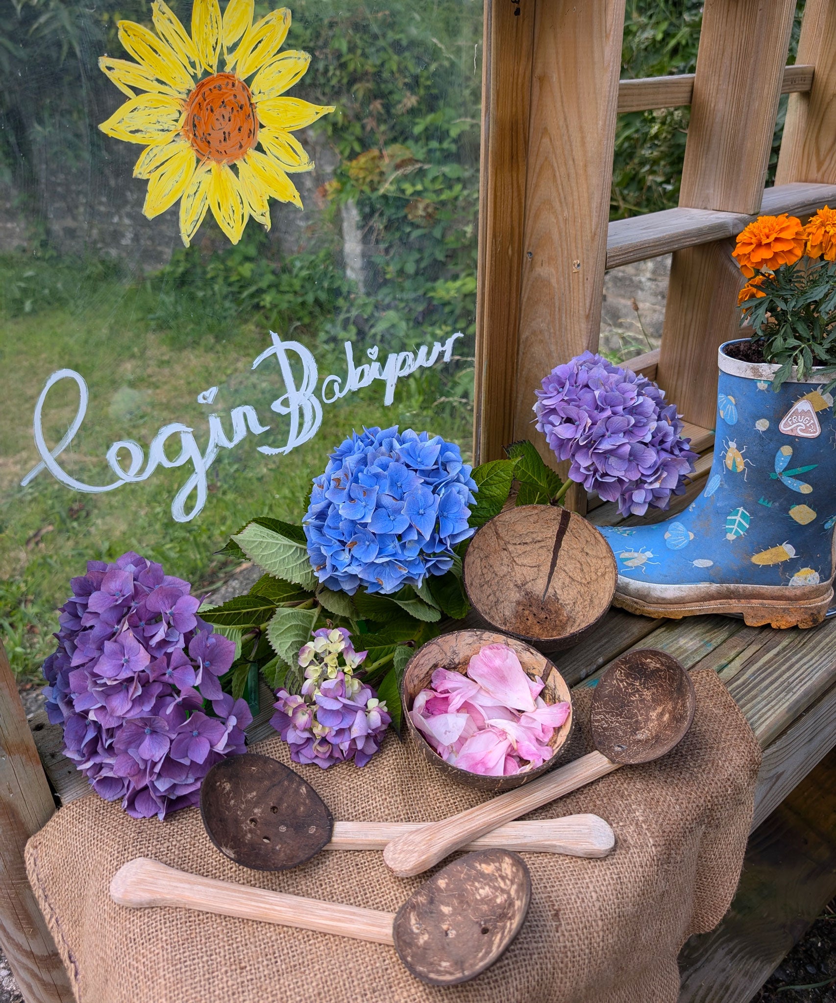 The Lanka Kade Coconut Shell Water Playset laid out on the surface of a mud kitchen outdoors. Blue and purple coloured hydrangeas have been placed behind them. A perspex board can be seen in the background with the words 'Cegin Babipur' which is welsh for Babipur Kitchen written with Kitpas crayons along with a drawing of a sunflower head.