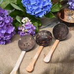 The three coconut shell utensils, slotted spoon, spoon and ladle spoon from the Lanka Kade water playset placed on jute material. Blue and purple hydrangeas can be seen in the background. 