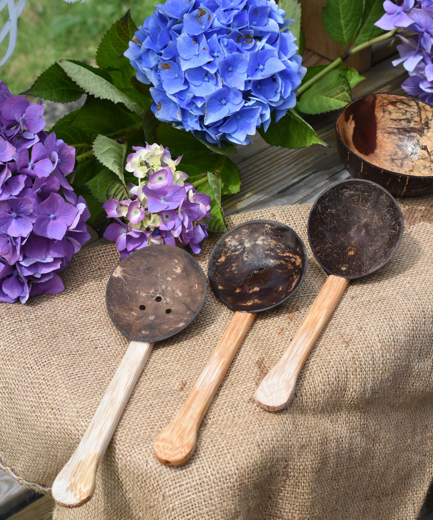 The three coconut shell utensils, slotted spoon, spoon and ladle spoon from the Lanka Kade water playset placed on jute material. Blue and purple hydrangeas can be seen in the background. 