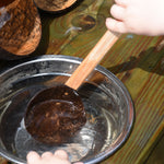 A close up of a child using the Lanka Kade coconut shell spoon utensil. The child is scooping water out of a small water bowl.