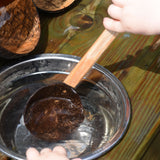 A close up of a child using the Lanka Kade coconut shell spoon utensil. The child is scooping water out of a small water bowl.