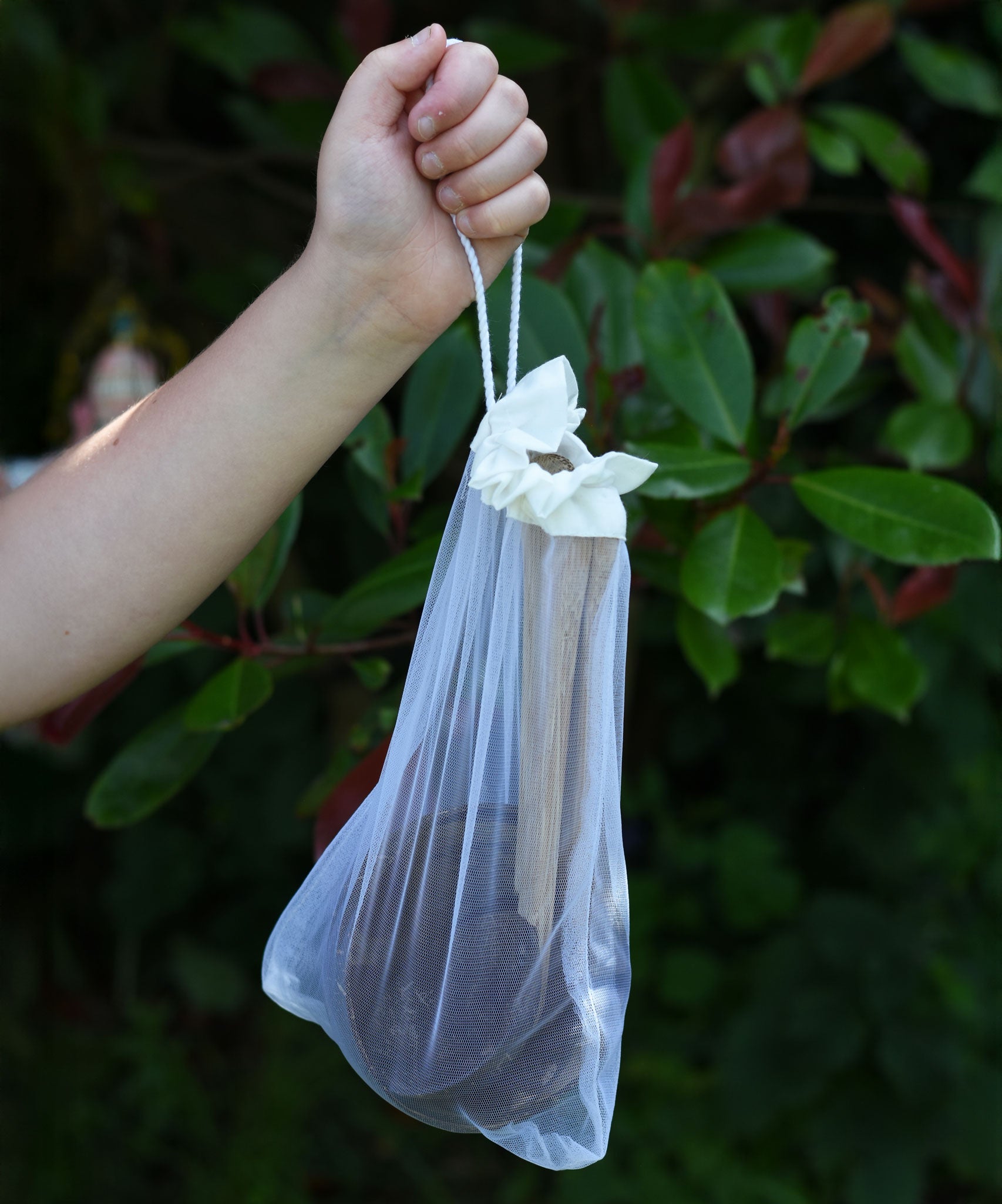 The Lanka Kade Coconut Shell Water Playset held up in a child's hand. The utensils and bowls have all been placed in a white mesh storage bag that come with the set.  