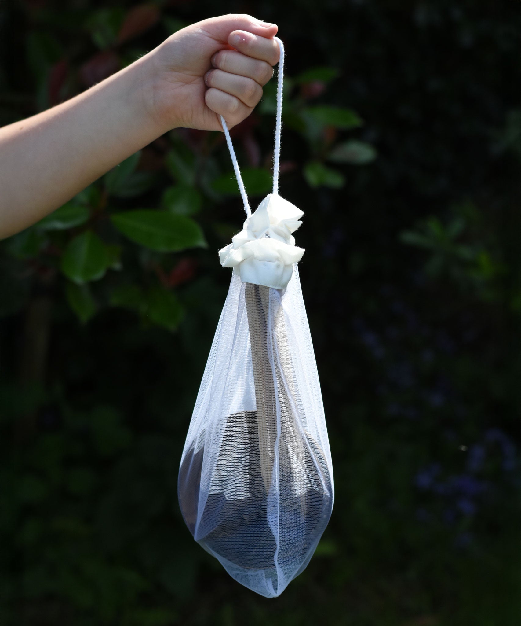 The Lanka Kade Coconut Shell Water Playset held up in a child's hand. The utensils and bowls have all been placed in a white mesh storage bag that come with the set.  