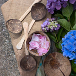 The utensils and bowls Lanka Kade Coconut Shell Water Playset placed on a jute material. There are pink coloured peony petal in the coconut bowl. Blue and purple coloured hydrangeas can be seen to the side. 